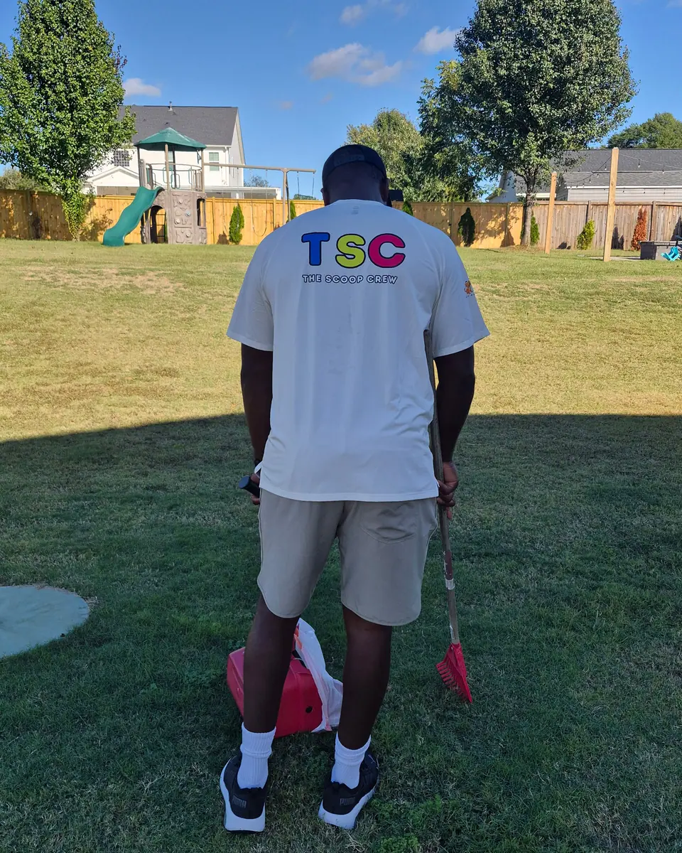 A Scoop Crew team member in a crew shirt, seen from behind, standing in a backyard with a rake and collection pan