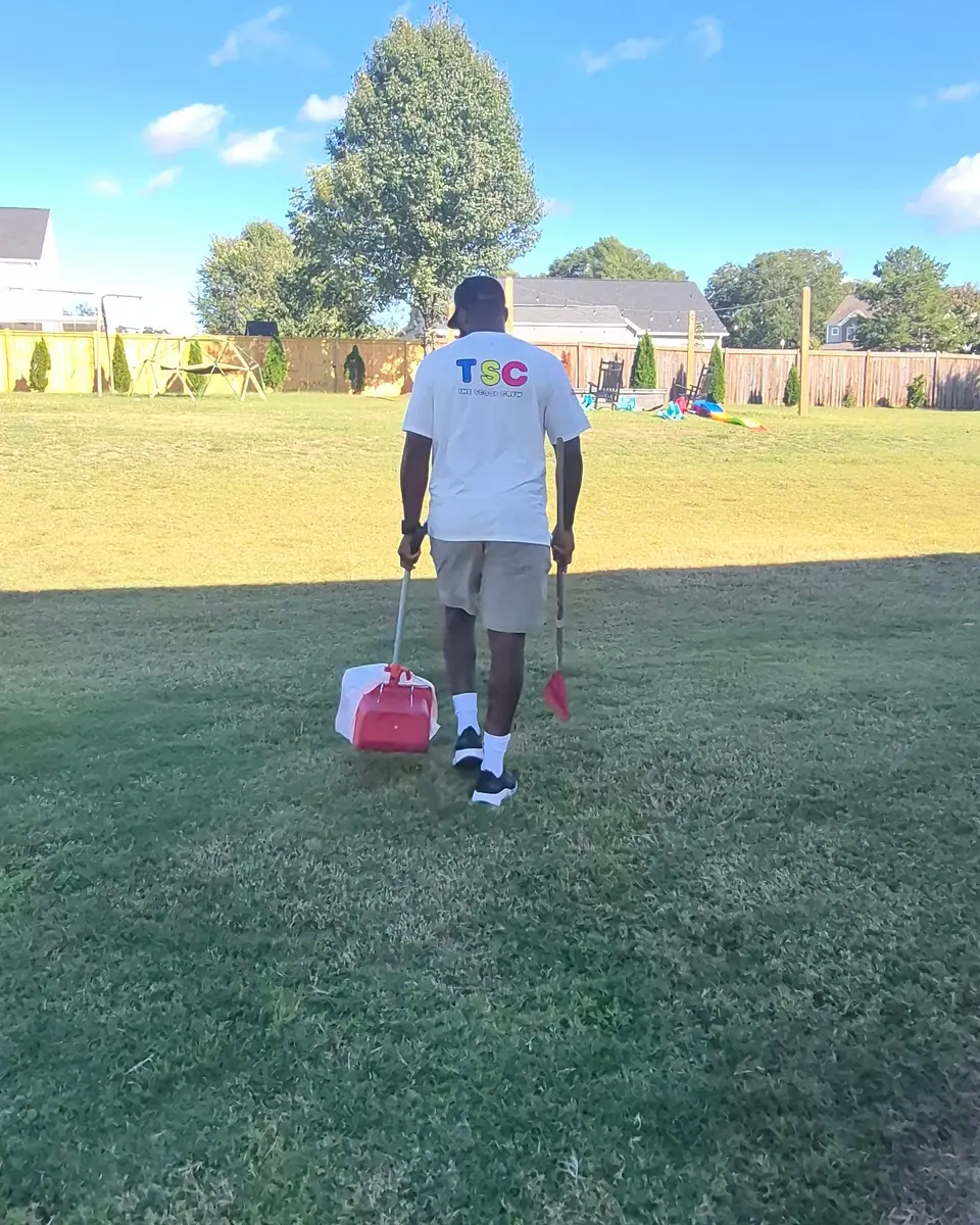 A Scoop Crew team member walking across a backyard carrying a rake and collection pan